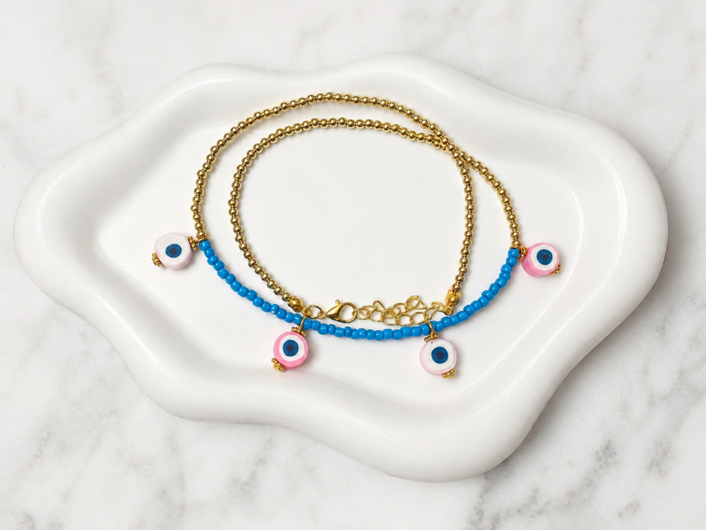 Handmade gold beaded necklace with turquoise seed beads and four pink evil eye charms displayed on a white decorative tray.
